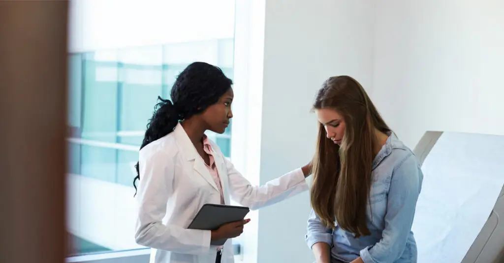 Here’s an optimized **alt text** for this image: Doctor in a white coat making a meaningful connection with a young woman sitting on an examination table in a medical office