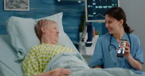 A nurse in blue scrubs smiling while showing a video call with a doctor to an elderly female patient lying in a hospital bed